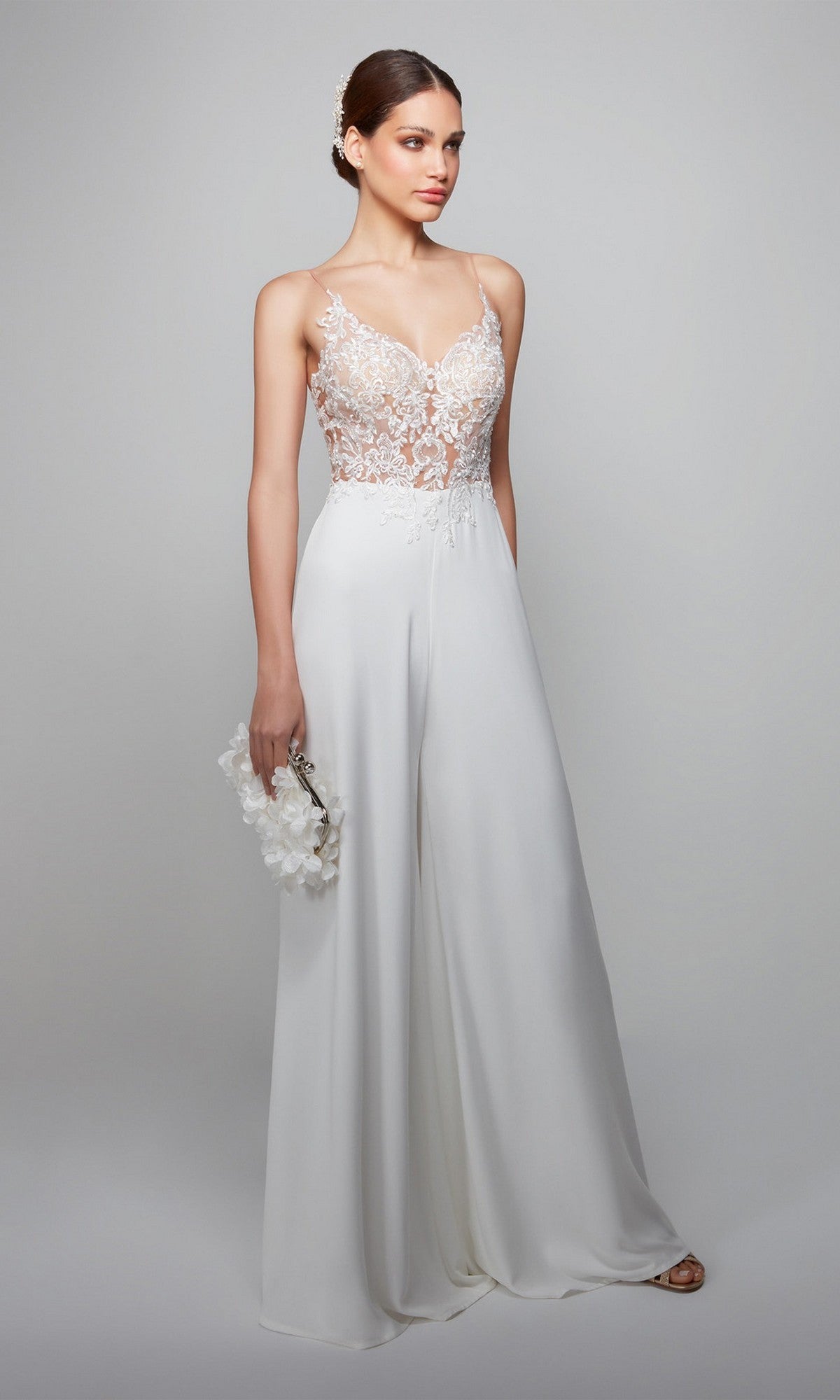 A woman wears the Long White Jumpsuit: Alyce 70017, which features a white lace bodice and wide-leg pants, holding a white floral clutch against a plain light background.